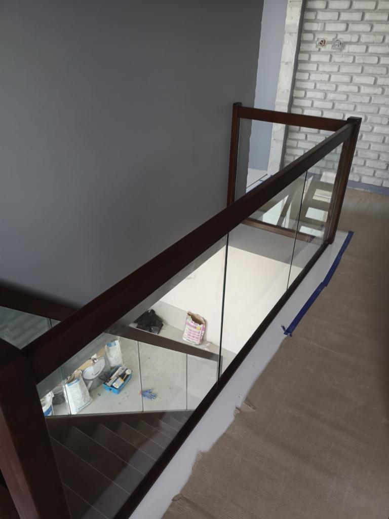 Nowoczesne schody z drewnianą balustradą i szklanym wypełnieniem, widok z góry, w tle ściana z białej cegły i szara ściana, widoczne ślady prac wykończeniowych na podłodze.