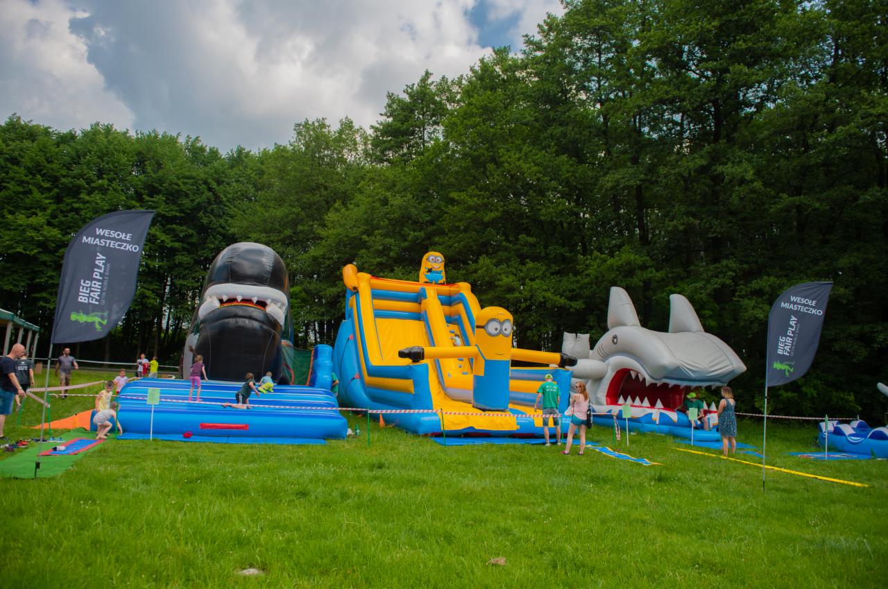 Rozległy park rozrywki na świeżym powietrzu z dmuchanymi atrakcjami: rekin, King Kong i zjeżdżalnia z Minionkami. W tle drzewa i flaga 'Wesołe Miasteczko'.