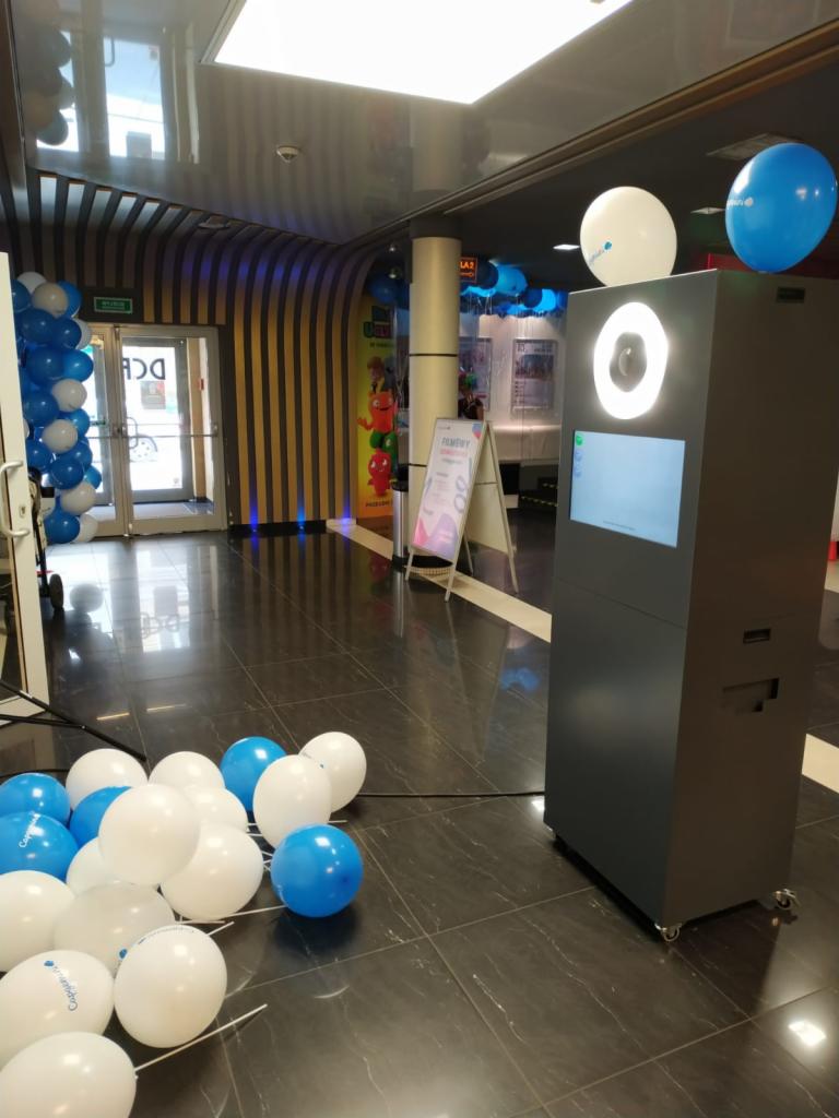 Ciemnoszara automat fotograficzny z białym światłem pierścieniowym, udekorowany białymi i niebieskimi balonami, ustawiony na marmurowej podłodze w centrum handlowym.