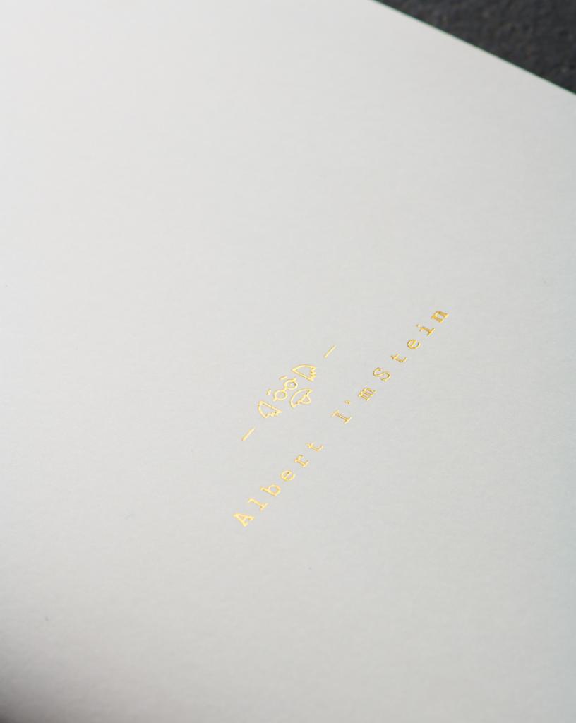 Biała kartka papieru z nadrukowanym złotym tekstem 'Albert I'm Stein' i małą grafiką powyżej, sfotografowana pod kątem.