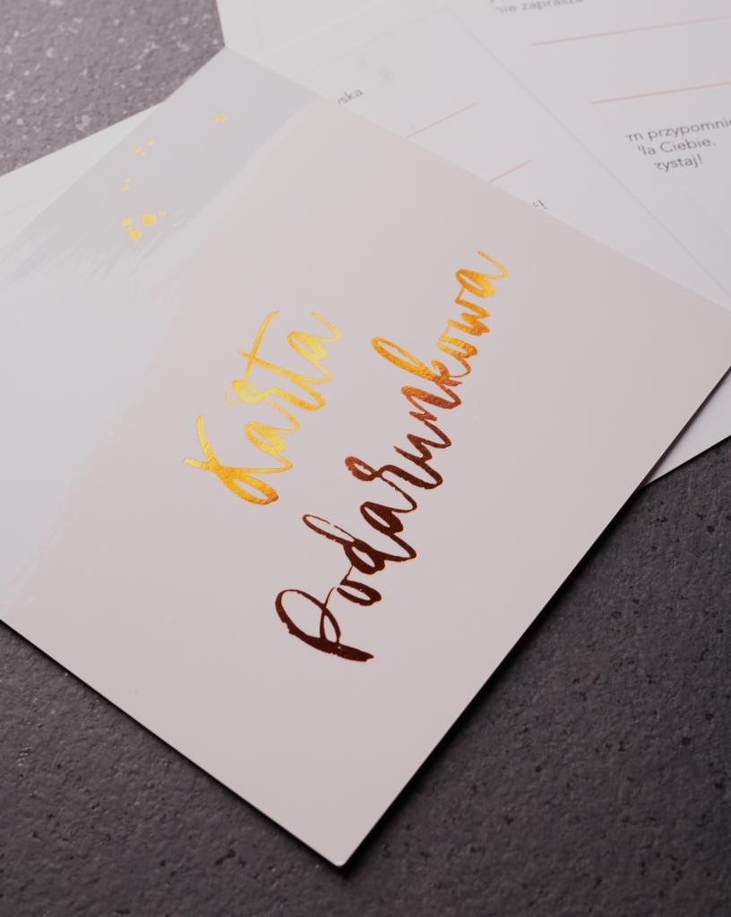 Karta podarunkowa z metalizowanym, złotym napisem kaligraficznym, leżąca na szarym tle, obok innych kart zaproszeń.