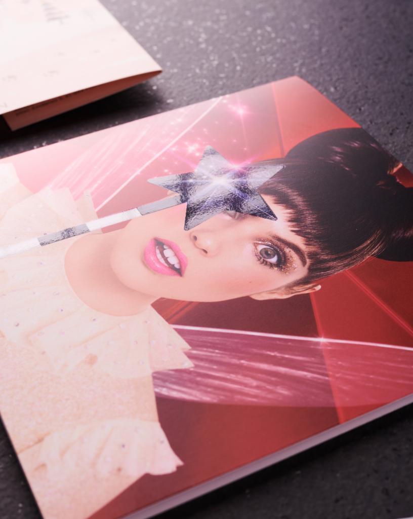 Katalog z okładką przedstawiającą portret kobiety z błyszczącą gwiazdą na oku, leżący na ciemnym blacie.