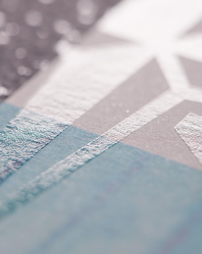 Zbliżenie na teksturowany papier z geometrycznym wzorem w odcieniach błękitu, szarości i srebra, z efektem połysku i delikatnym ziarnem.