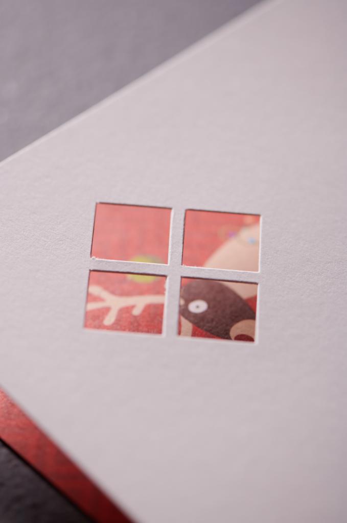 Makro zbliżenie na kartkę z wyciętym motywem okna, przez które widać fragment ilustracji świątecznej z reniferem, na szarym tle.