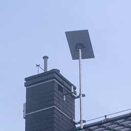 Satelis Marcin Maniecki - Płaska antena zamontowana na metalowym maszcie, przymocowanym do komina na dachu pokrytym ciemną dachówką. Widok z dołu, pochmurne niebo w tle.