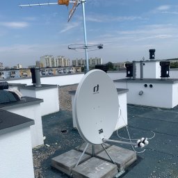 Satelis Marcin Maniecki - Antena satelitarna Invertro i antena naziemna Yagi-Uda zamontowane na płaskim dachu budynku mieszkalnego, widok na miasto w tle, błękitne niebo.