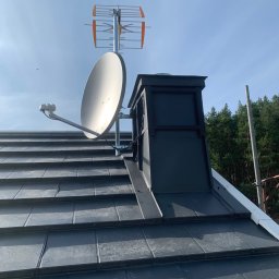 Satelis Marcin Maniecki - Antena satelitarna i naziemna zamontowane na dachu z blachy na rąbek stojący, komin w obudowie z blachy, las w tle, błękitne niebo.