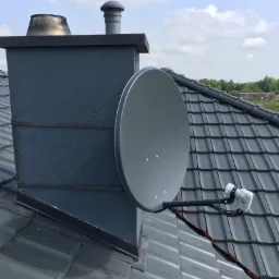 Szara antena satelitarna zamontowana na kominie obok grafitowego dachu z dachówki ceramicznej, widok z góry na tle błękitnego nieba z chmurami.