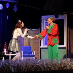 Scena z przedstawienia dla dzieci: kobieta w srebrnej spódnicy i mężczyzna przebrany za elfa śpiewają do mikrofonów, trzymając się za ręce. W tle choinka i prezenty.