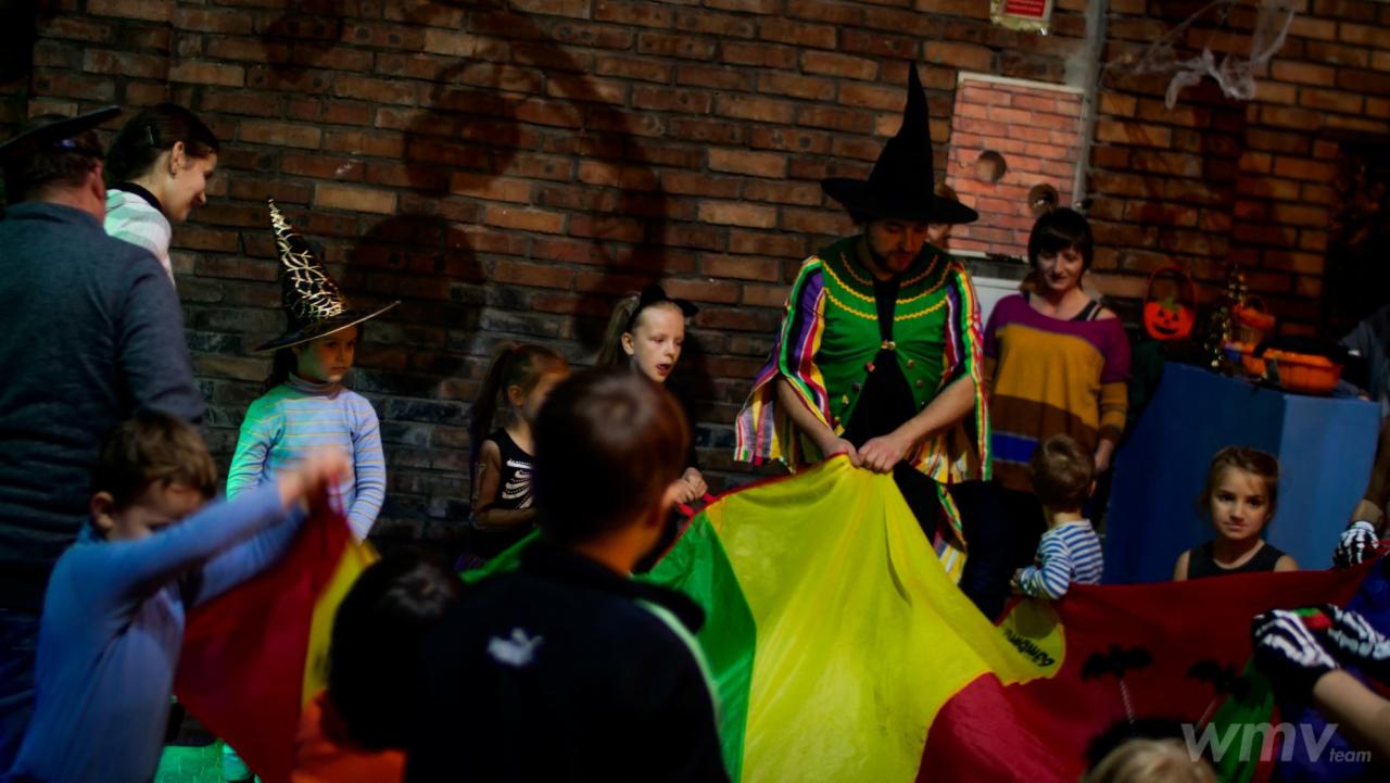 Dzieci w kostiumach bawią się kolorową chustą animacyjną podczas imprezy Halloween, prowadzonej przez mężczyznę w stroju czarownika na tle ceglanej ściany z dekoracjami.