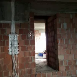 Rozdzielnica elektryczna z podłączonymi przewodami na surowej ścianie z czerwonej cegły w trakcie prac instalacyjnych, widoczne kable prowadzone pod sufitem i paleta na podłodze.