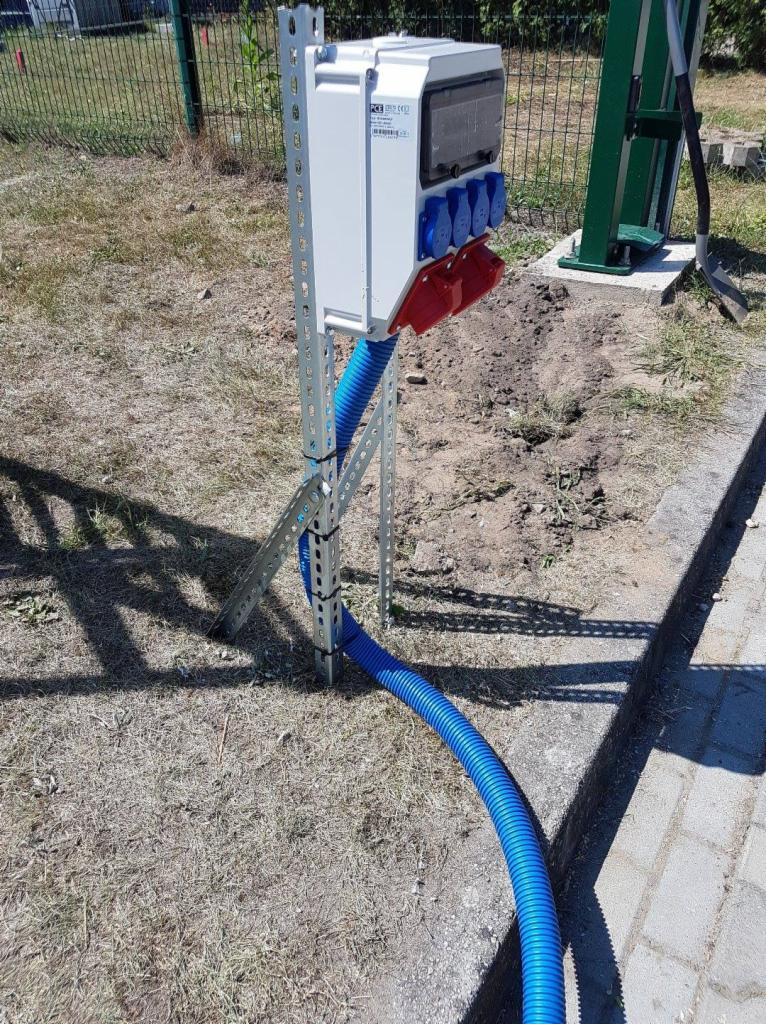 Zewnętrzna skrzynka elektryczna z gniazdami siłowymi (czerwone i niebieskie) zamontowana na metalowym stelażu, z niebieskim karbowanym peszlem prowadzącym kable w ziemi.