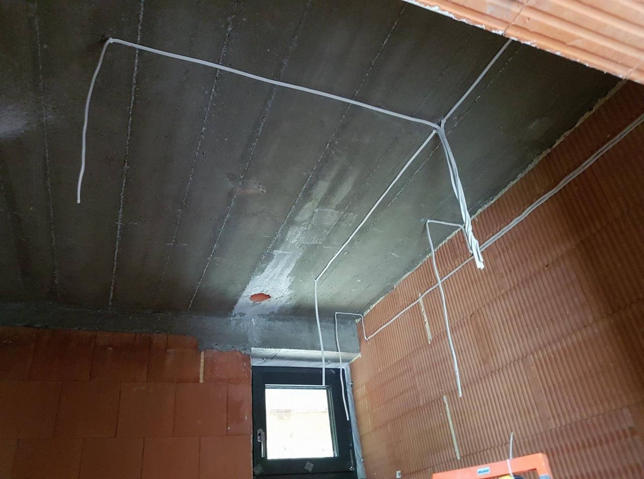 Surowy betonowy strop z widoczną instalacją elektryczną w białych peszlach, ściany z czerwonej cegły, okno w tle. Widać etap prac instalacyjnych w nowym budownictwie.