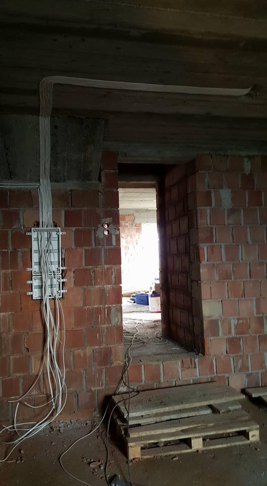 Rozdzielnica elektryczna z podłączonymi przewodami na surowej ścianie z czerwonej cegły w trakcie prac instalacyjnych, widoczne kable prowadzone pod sufitem i paleta na podłodze.