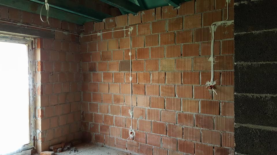 Surowa ściana z czerwonej cegły dziurawki z częściowo poprowadzoną instalacją elektryczną; widoczne kable i puszki elektryczne, obok fragment ściany z czarnego betonu komórkowego, okno w lewym...