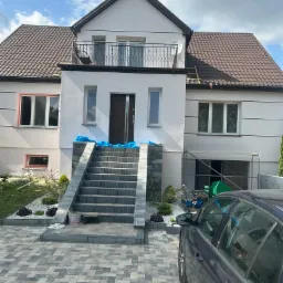 Dom z balkonem w trakcie prac elewacyjnych: zabezpieczone okna folią, widoczne rusztowania, schody wejściowe z kamienia. Częściowo otynkowana fasada.