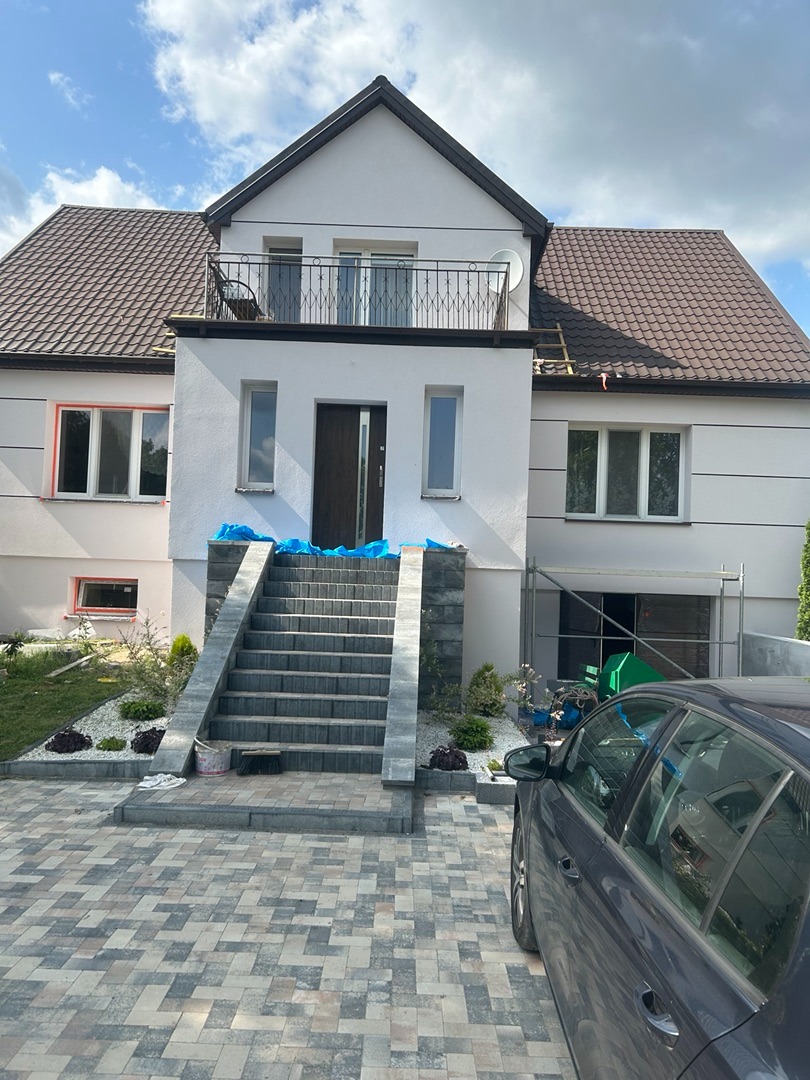 Dom z balkonem w trakcie prac elewacyjnych: zabezpieczone okna folią, widoczne rusztowania, schody wejściowe z kamienia. Częściowo otynkowana fasada.