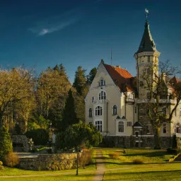 Elegancki, kremowy budynek z wieżą i czerwonym dachem otoczony zielonym trawnikiem i drzewami we Włokach, przypominający pałac lub luksusowy hotel.