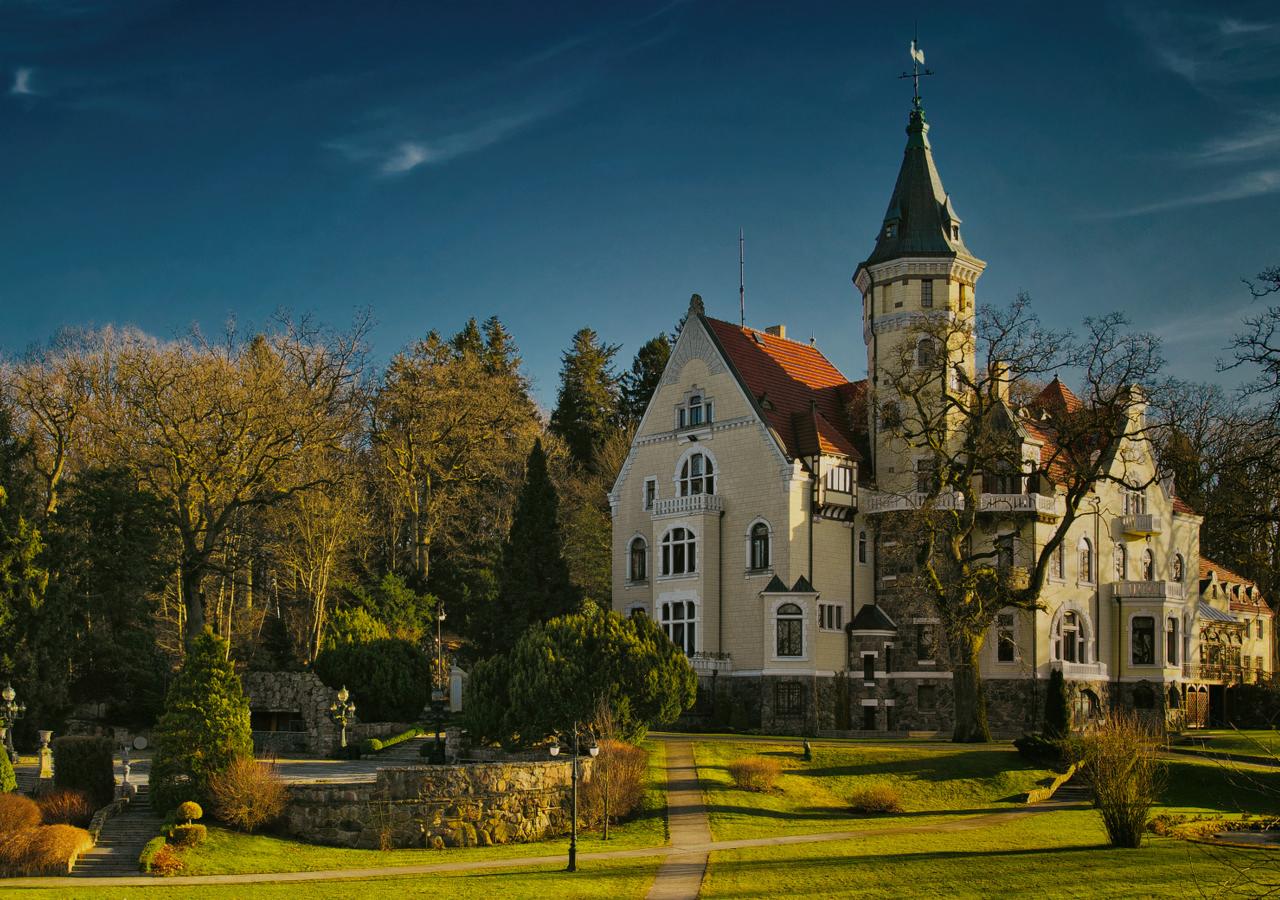 Elegancki, kremowy budynek z wieżą i czerwonym dachem otoczony zielonym trawnikiem i drzewami we Włokach, przypominający pałac lub luksusowy hotel.