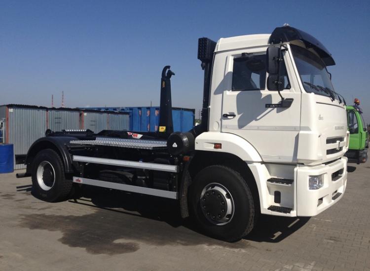 Biała ciężarówka marki Kamaz z systemem hakowym do transportu kontenerów, zaparkowana na placu z widocznymi kontenerami w tle.