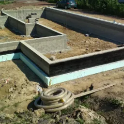 Wykopane fundamenty domu z bloczków betonowych, zabezpieczone izolacją termiczną w kolorze jasnoniebieskim oraz czarną hydroizolacją. W tle widoczny zaparkowany samochód.