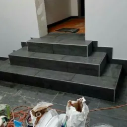 Wyłożone ciemnymi płytkami schody wejściowe do pomieszczenia, z widocznymi śladami prac wykończeniowych i narzędziami na podłodze.