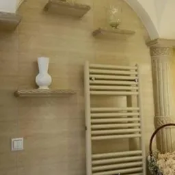 Beżowa łazienka z grzejnikiem drabinkowym, trzema półkami w niszy zwieńczonej łukiem, na których stoją dekoracje: złota maska, szklany wazon i biały wazon ceramiczny.