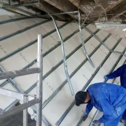 Montaż płyt gipsowo-kartonowych na metalowym ruszcie pod skosem, widoczne fragmenty starego stropu i ocieplenie, pracownik w niebieskim kombinezonie przy pracy.