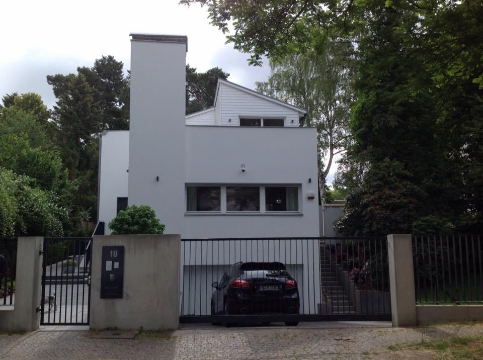 Nowoczesny dom z płaskim dachem i wysokim kominem, czarna brama wjazdowa z numerem posesji, zaparkowany czarny samochód.