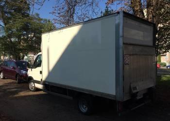 Biały samochód dostawczy z zabudową kontenerową zaparkowany na poboczu, widoczny z boku, z drzewami w tle.