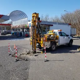 Żółta wiertnica zamontowana na białym pick-upie wykonuje odwiert w pobliżu stacji benzynowej, teren zabezpieczony pachołkami i taśmą.
