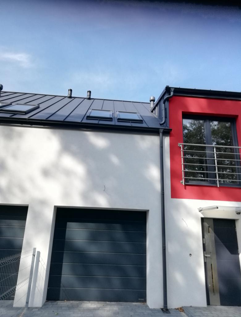 Nowoczesny dom z ciemnoszarym dachem blaszanym z oknami dachowymi i rynnami w kolorze antracytu. Część elewacji biała, część czerwona. Widok z dołu.