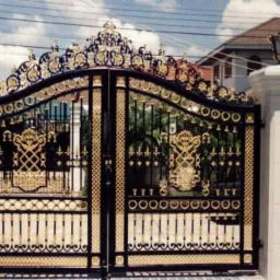 Ozdobna, dwuskrzydłowa brama wjazdowa z elementami kutymi w kolorze złotym, osadzona na masywnych, białych filarach z lampami.