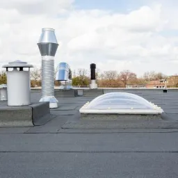 Czarny, płaski dach pokryty papą z kominami wentylacyjnymi, świetlikiem dachowym i obróbkami blacharskimi na tle błękitnego nieba z chmurami.