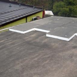 Kramar - Usługi Remontowo-Budowlane - Płaski dach pokryty papą z fragmentami termoizolacji z białym obrzeżem, widok z góry na fragment dachu budynku z żółtą ścianą i brązowym dachem w tle.