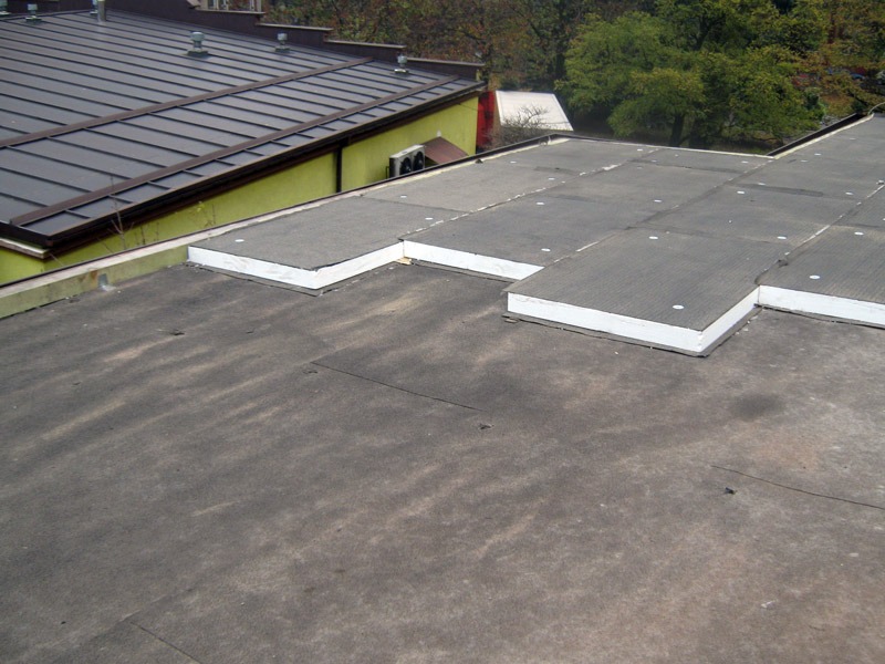 Płaski dach pokryty papą z fragmentami termoizolacji z białym obrzeżem, widok z góry na fragment dachu budynku z żółtą ścianą i brązowym dachem w tle.