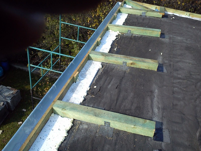 Dach pokryty papą z drewnianą konstrukcją mocowaną do podłoża, widoczna piana uszczelniająca i fragment rusztowania.