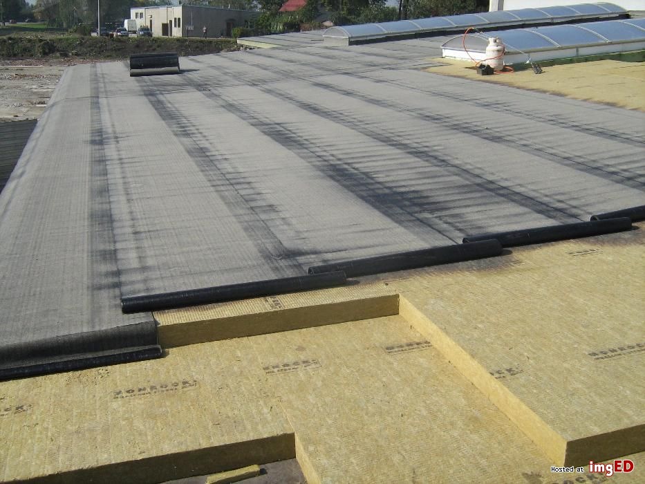 Dach płaski w trakcie izolacji: wełna mineralna ułożona warstwowo, częściowo pokryta papą termozgrzewalną, widoczne rolki papy i butla gazowa w tle.