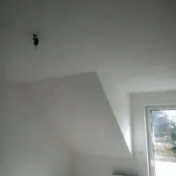 Poddasze po remoncie z gładkimi, białymi ścianami i skośnym sufitem, widoczny grzejnik i okno z widokiem na zewnątrz, żarówka na kablu zwisająca z sufitu.