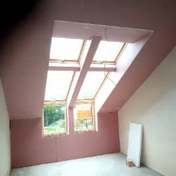 Poddasze w trakcie wykańczania: widoczne okna dachowe, płyty gipsowo-kartonowe w kolorze r&oacute;żowym na skosach, oraz fragment ściany pokrytej szpachlą. Widoczny przew&oacute;d wentylacyjny.