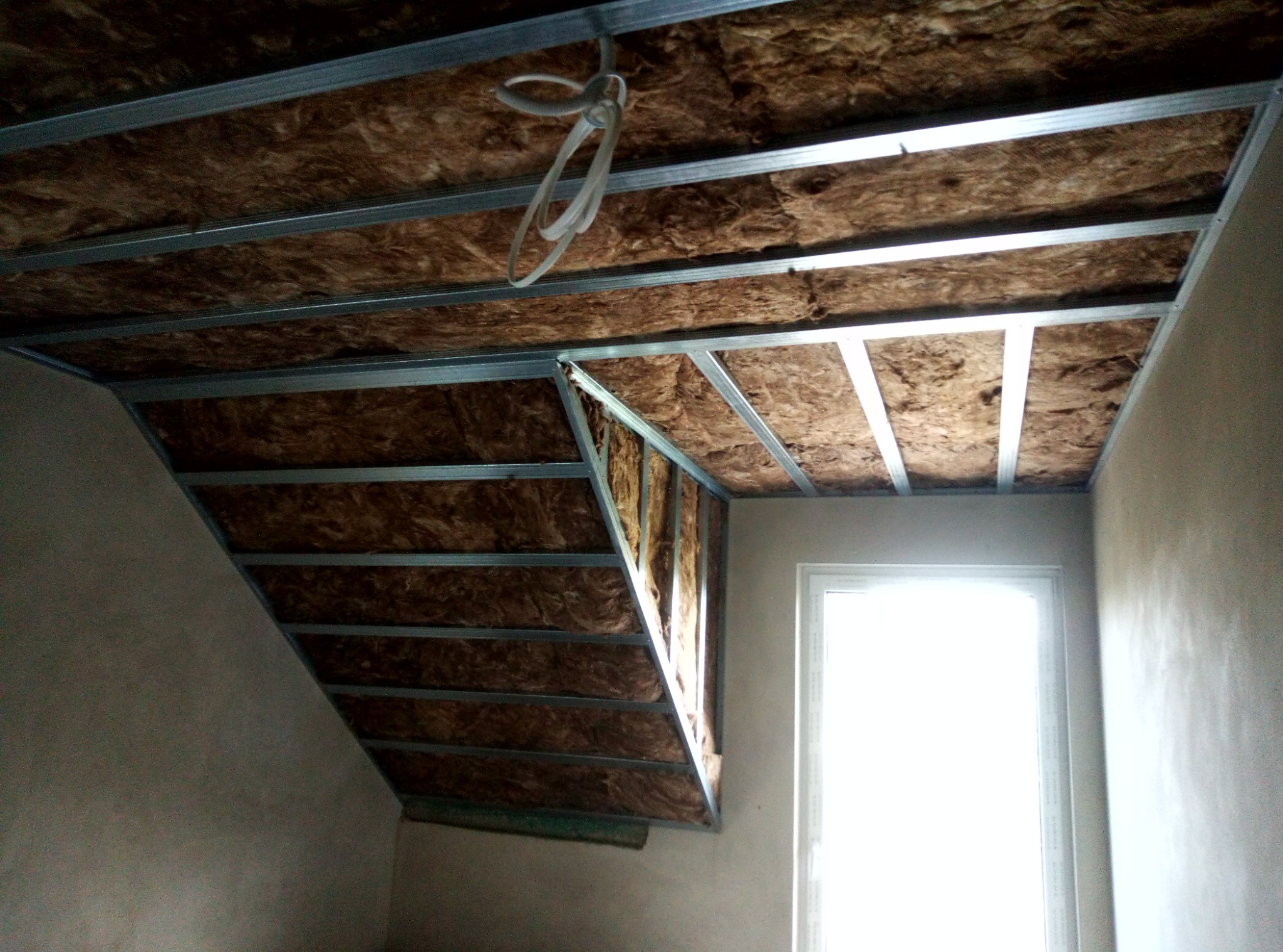 Skośne poddasze w trakcie ocieplania wełną mineralną, widoczna metalowa konstrukcja nośna, fragment okna w tle.