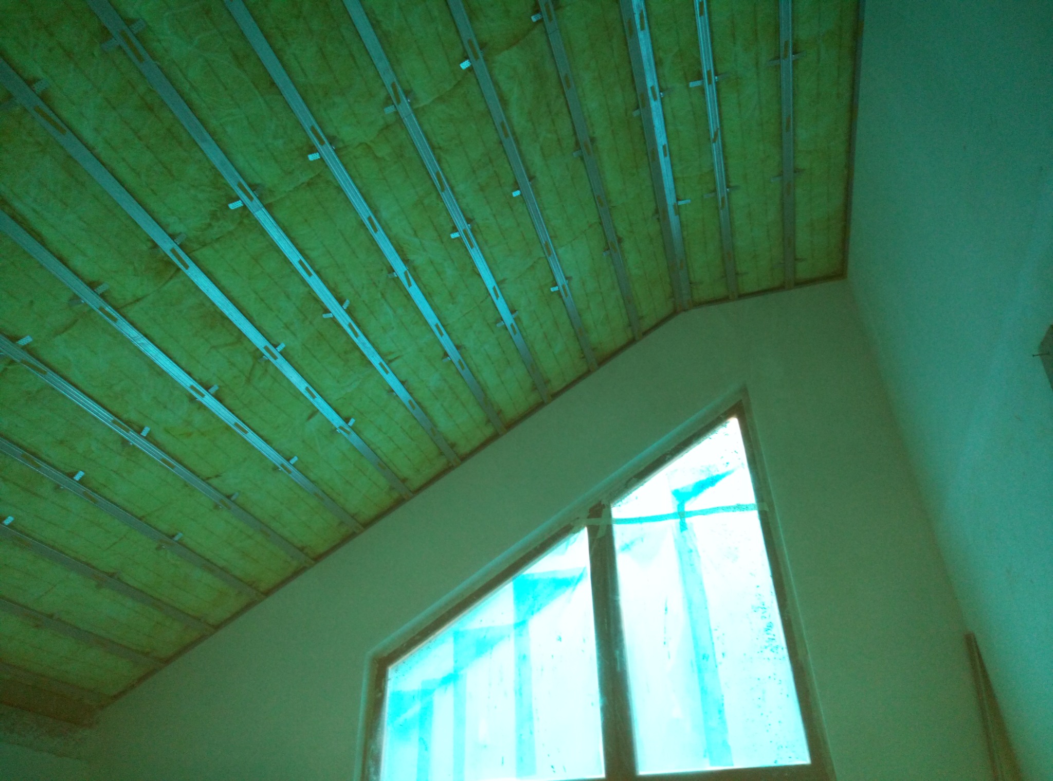 Skośne poddasze w trakcie ocieplania, widoczne wełna mineralna między metalowymi profilami, okno trójkątne zabezpieczone folią.