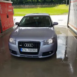 Szary samochód Audi A3 po umyciu w samoobsługowej myjni, widoczne krople wody na karoserii i mokra posadzka. W tle zieleń i stanowiska myjące.