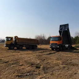 Dwa samochody ciężarowe na polu: ż&oacute;łty wywrotka i pomarańczowy wywrotka z uniesioną skrzynią ładunkową, wysypujący materiał na ziemię.