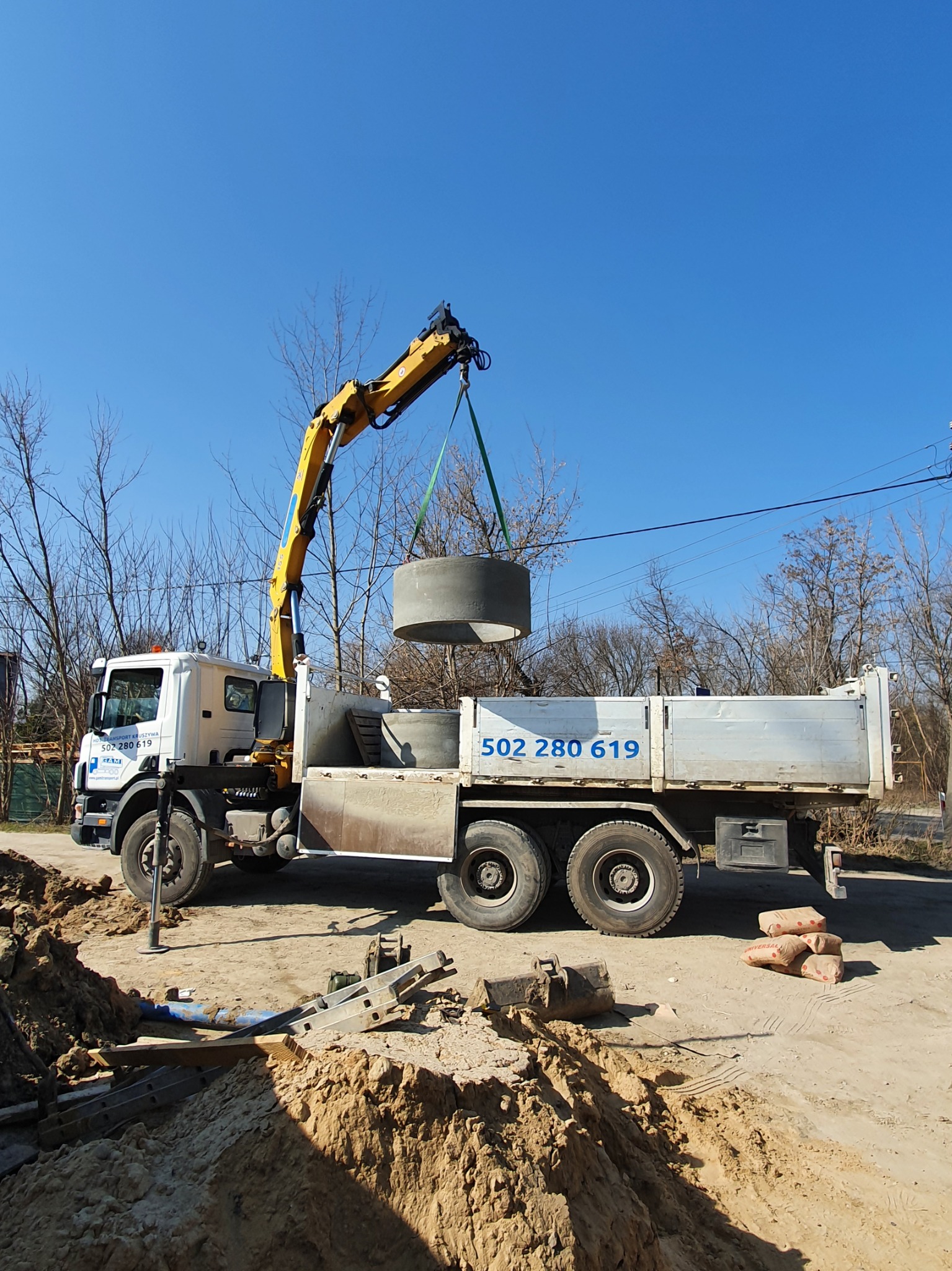 Ciężarówka z dźwigiem podnosi betonowy krąg szamba na tle wykopu i błękitnego nieba. Na burcie ciężarówki widoczny numer telefonu 502-280-619.