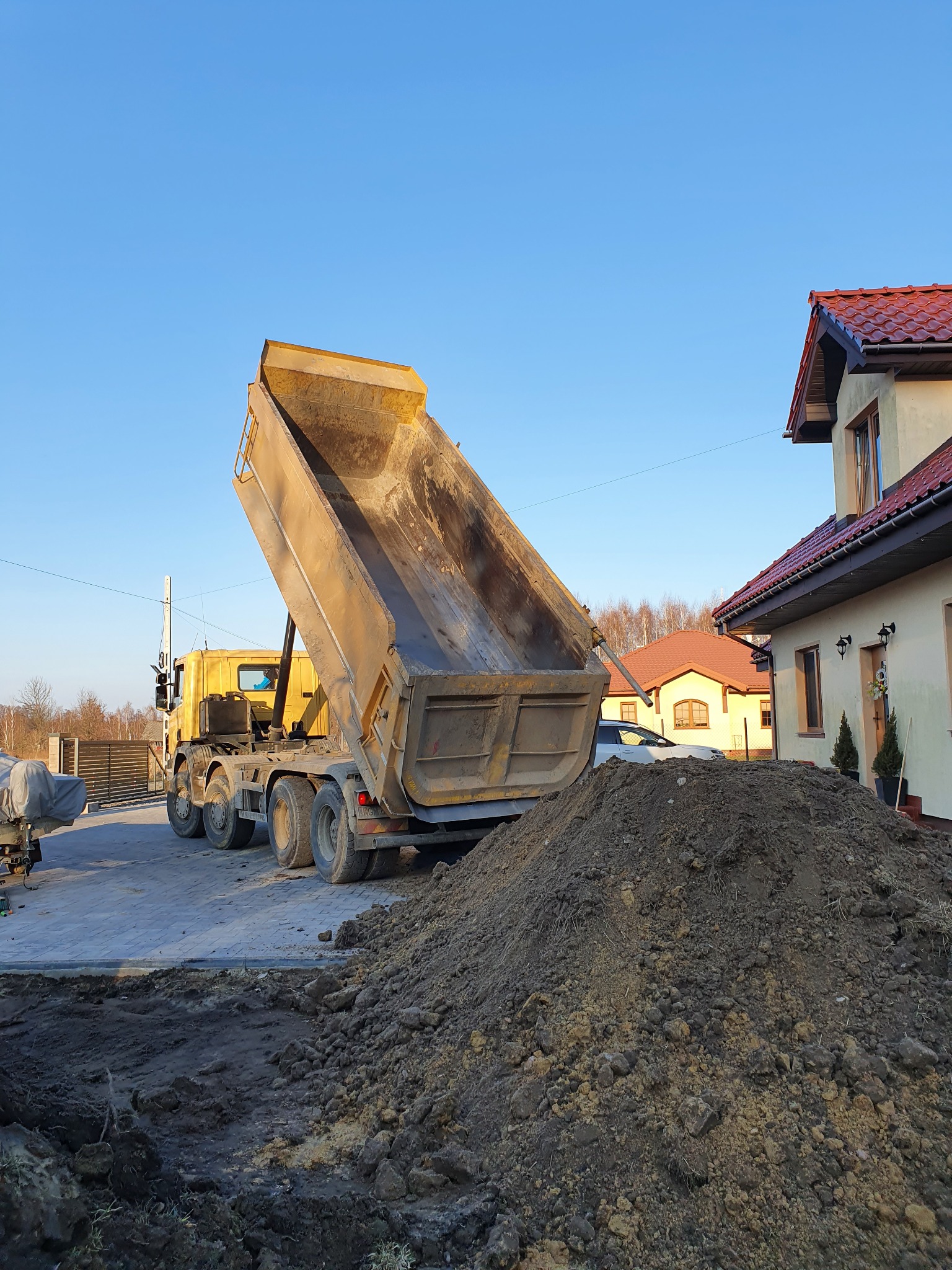 Żółta wywrotka z podniesioną skrzynią wyładowuje ziemię na placu budowy przed domem z czerwoną dachówką.