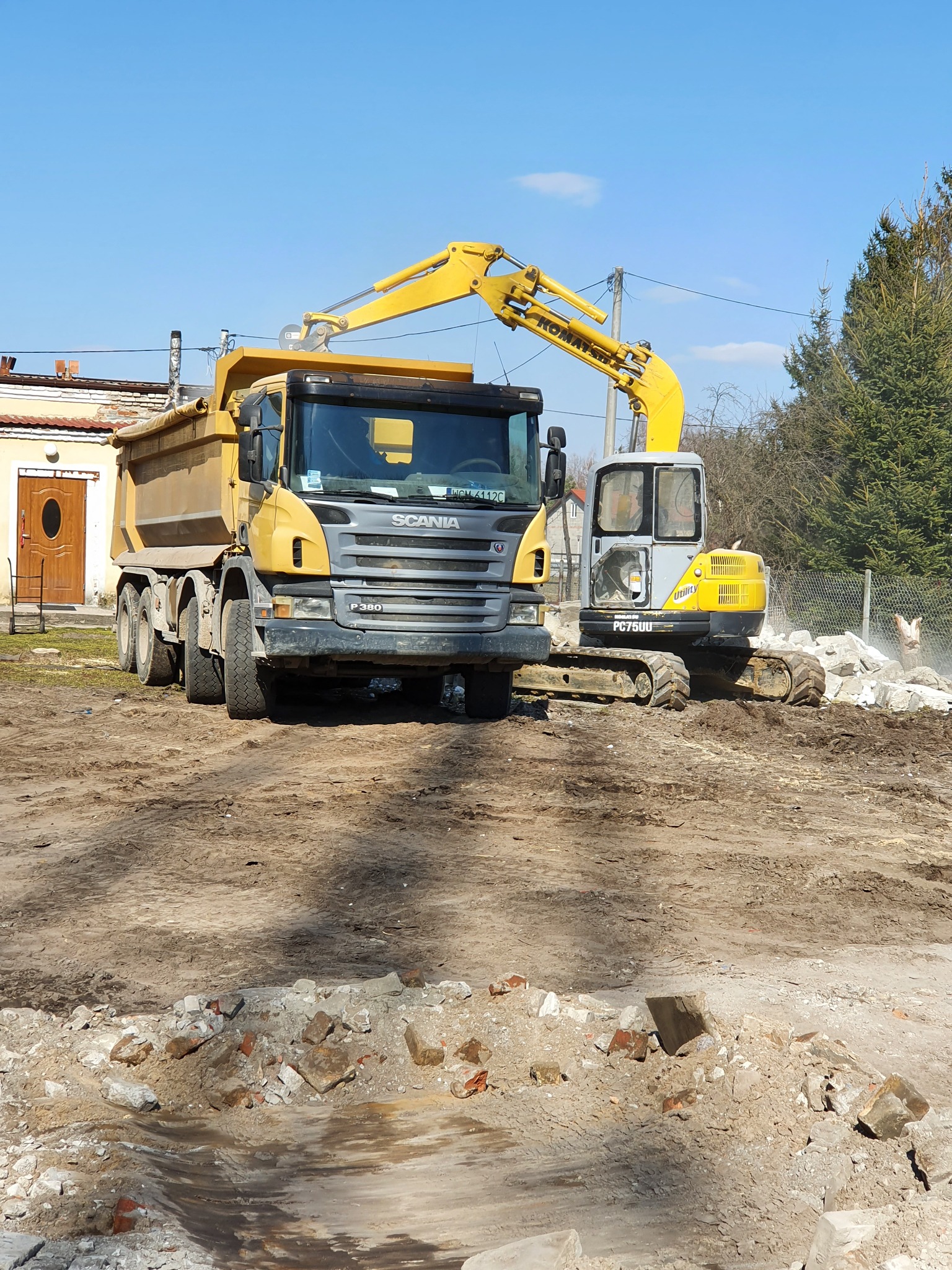 Żółta wywrotka Scania P380 i koparka Komatsu PC75UU na placu budowy z gruzem, widok z przodu, błękitne niebo w tle.