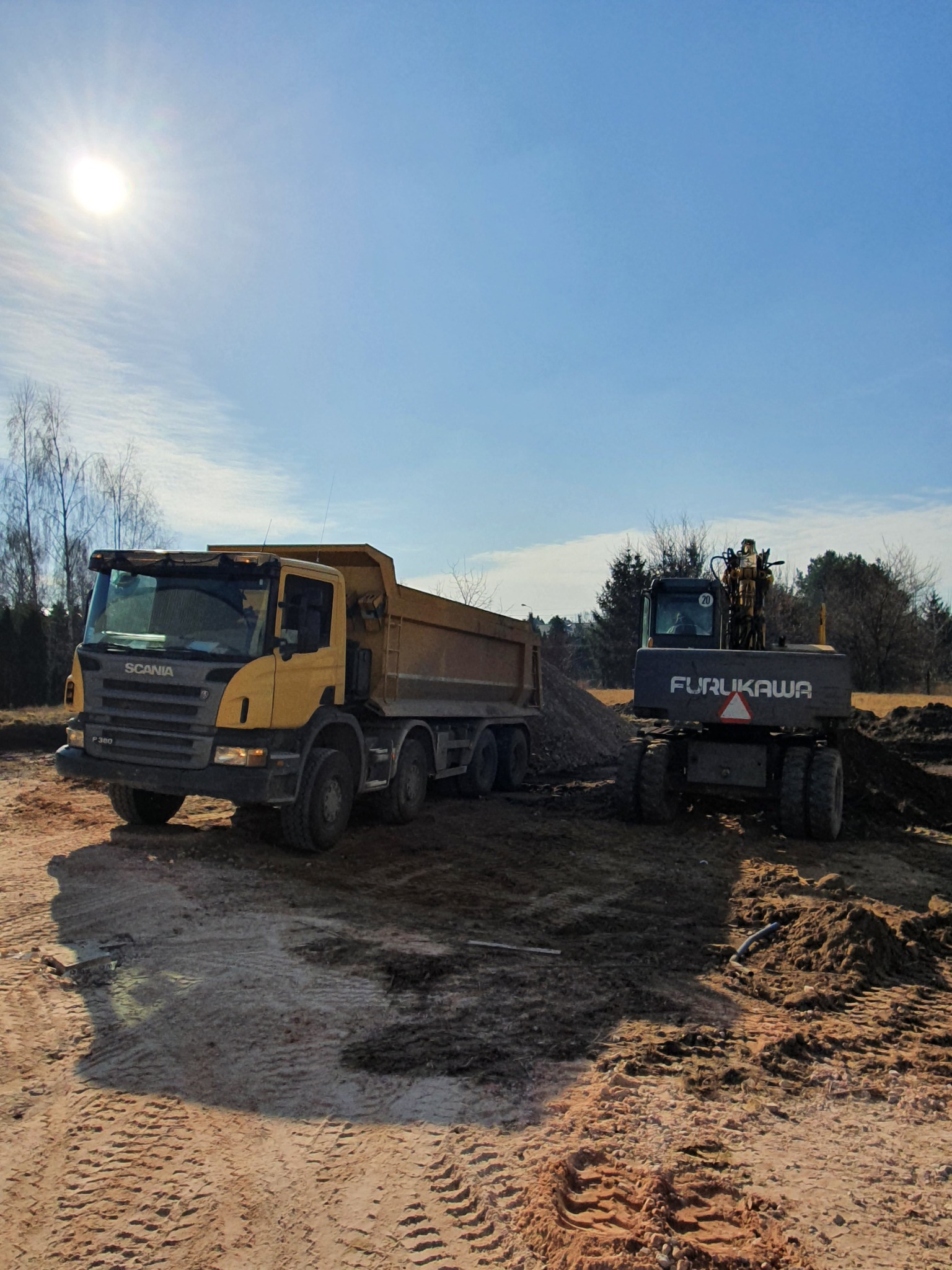 Żółta wywrotka Scania i szara koparka Furukawa na placu budowy, widoczne ślady opon na ziemi, w tle drzewa i błękitne niebo z promieniami słońca.