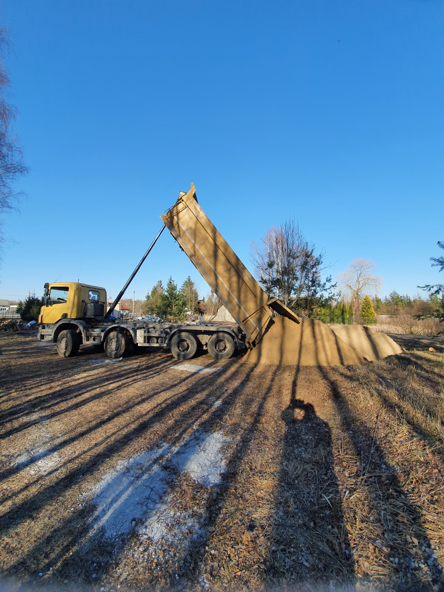 Żółta wywrotka w trakcie wysypywania ładunku na placu, widoczny cień fotografa.