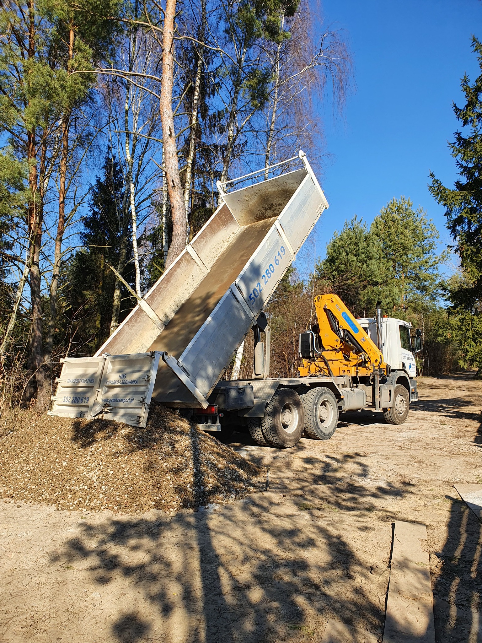 Wyładowany z wywrotki żwir na terenie z drzewami w tle, widoczny numer telefonu na burcie
